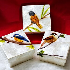 May include: Three small white boxes with different bird designs. The boxes are made of paper mache and have a textured surface. The birds are painted in bright colors and are perched on branches. The boxes are perfect for storing small items or as decorative accents.