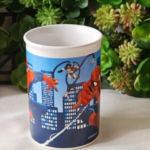 Puede incluir: Una taza de cer&aacute;mica blanca con un dise&ntilde;o azul, rojo y negro con personajes de Spider-Man y el texto "&copy;MARVEL".