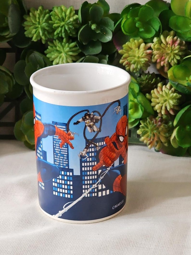 Puede incluir: Una taza de cer&aacute;mica blanca con un dise&ntilde;o azul, rojo y negro con personajes de Spider-Man y el texto "&copy;MARVEL".