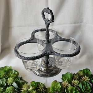 May include: A three-tiered metal serving caddy featuring a dark, ornate, matte-finished frame with intricate scrollwork details. The stand includes a central handle and holds three clear, ribbed glass tumblers arranged in a circular formation.
