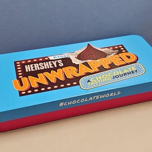 Può includere: Una scatola di caramelle rettangolare Hershey's Unwrapped con coperchio blu e base rosa. Il coperchio presenta il logo Hershey's, una grafica di bacio al cioccolato e il testo "A Chocolate Tasting Journey". L'hashtag #chocolateworld è visibile.