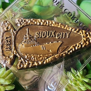 Puede incluir: Una moneda de desafío de metal de color dorado en forma de punta de flecha con las palabras "Sioux City" y "LCS 11" en ella. La moneda presenta un barco con el número "11" en el lateral.