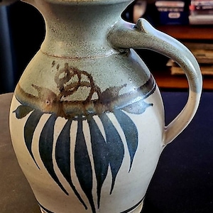 Può includere: Brocca in ceramica con smalto verde chiaro e beige. La brocca presenta un manico curvo ed è decorata con motivi a forma di foglie blu scuro e accenti marroni. La brocca misura circa 20 cm di altezza.