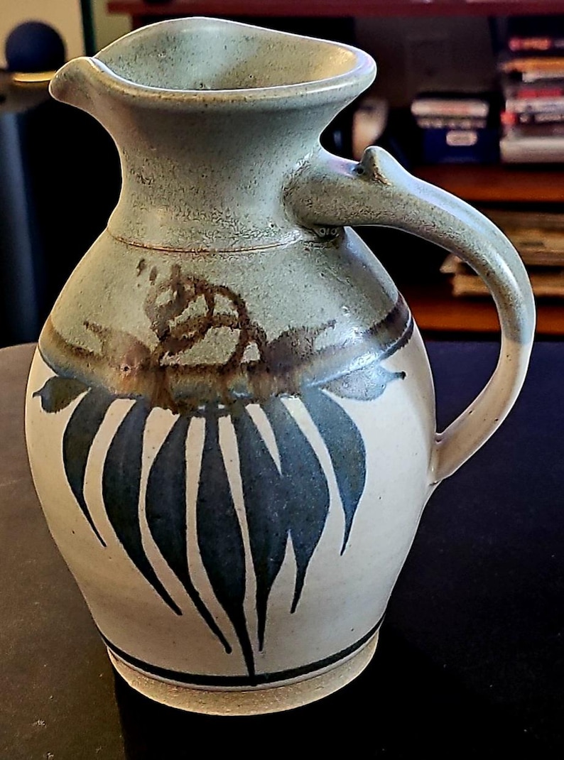 Op de afbeelding: Een keramische kan met een lichtgroen en beige glazuur. De kan heeft een gebogen handvat en is versierd met donkerblauwe bladachtige patronen en bruine accenten. De kan is ongeveer 20 cm hoog.