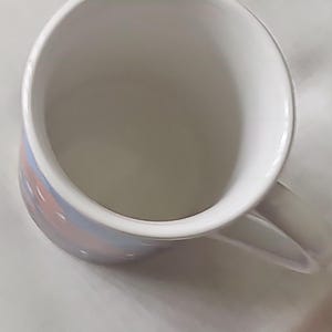Puede incluir: Una taza de cer&aacute;mica blanca con un dise&ntilde;o rosa y azul. La taza tiene un asa y est&aacute; vac&iacute;a.