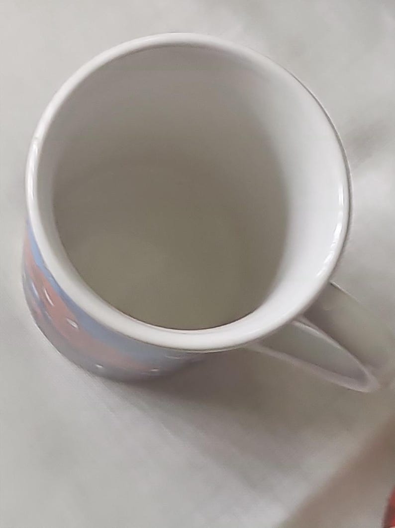 Puede incluir: Una taza de cer&aacute;mica blanca con un dise&ntilde;o rosa y azul. La taza tiene un asa y est&aacute; vac&iacute;a.