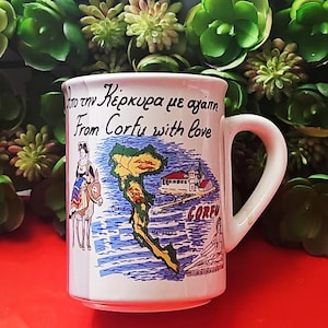 May include: White ceramic mug with a handle, featuring a map of Corfu, Greece, and the text "From Corfu with love." The mug has illustrations of a person on a donkey and a church. The mug is surrounded by green plants.