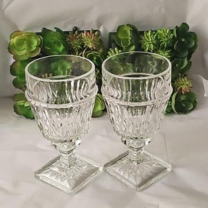May include: Two clear, vintage-style goblets with textured, vertical ribbing on the bowl and a square base. The stem is short and features a decorative pattern. The goblets are made of glass and are suitable for serving beverages.