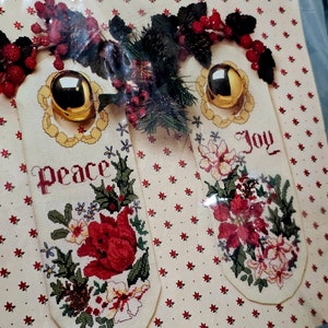 以下が含まれることがあります： 赤いポインセチアと緑の葉を特徴とする2つのクリスマスオーナメントのクロスステッチパターン。オーナメントには「Peace」と「Joy」のラベルが付けられています。