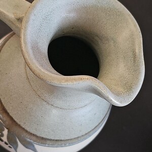Op de afbeelding: Een keramische kan met een brede opening en een gebogen tuit. De kan heeft een gedempte groene kleur met een textuur en een decoratief bladpatroon rond de basis. De handgreep is gebogen.