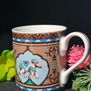Könnte beinhalten: Eine braune Keramiktasse mit einem weißen Henkel. Die Tasse hat ein blaues, braunes und weißes Muster mit einem Elefanten, der einen blauen und grünen Sattel trägt.