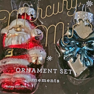 May include: A set of three glass Christmas ornaments. One ornament features a Santa Claus figure with a white beard and a red suit. Another ornament is a silver gift box with a blue bow. The ornaments are packaged in a box with the text "Mercury's Mirth Ornament Set" and "ornaments".