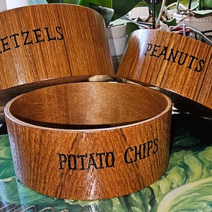 以下が含まれることがあります： 蓋付きの木製のボウルが3つあり、それぞれ異なるスナックのラベルが貼られています。「プレッツェル」、「ピーナッツ」、「ポテトチップス」。
