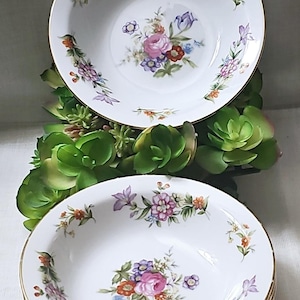 Op de afbeelding: Set van drie witte keramische kommen met een gouden rand. Elke kom heeft een bloemenmotief met roze, paarse en oranje bloemen.