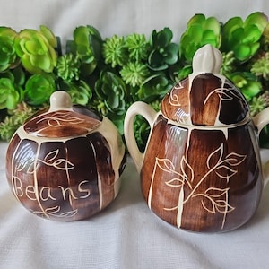 May include: Two ceramic kitchen canisters with lids. One is round and labeled "Beans" in white script. The other is a sugar bowl with handles. Both are brown and cream with leaf designs.