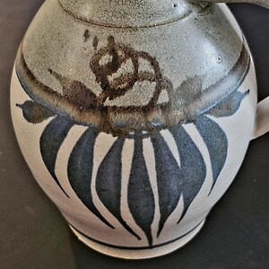 Op de afbeelding: Een keramische kan met een cr&egrave;mekleurige basis en een grijs bovenstuk. De kan heeft een donkerblauw en wit bladerenontwerp en een bruin bloemenaccent. Het heeft een handvat en een tuit.