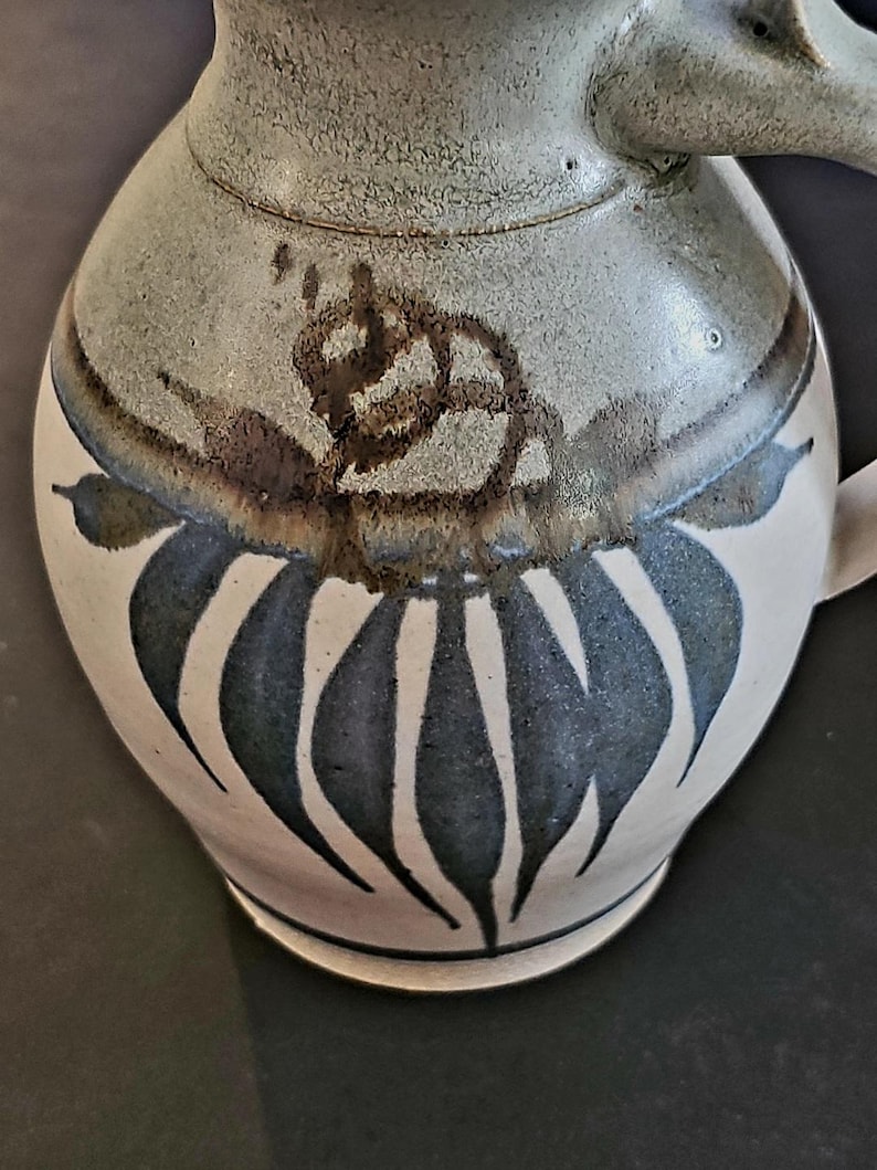 Op de afbeelding: Een keramische kan met een cr&egrave;mekleurige basis en een grijs bovenstuk. De kan heeft een donkerblauw en wit bladerenontwerp en een bruin bloemenaccent. Het heeft een handvat en een tuit.