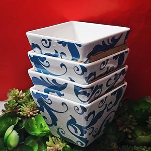以下が含まれることがあります： 青いフローラルデザインの白い正方形のセラミックボウルが4つ積み重なっています。ボウルは互いの上に積み重ねられ、背景は赤色です。ボウルの周りには緑色の植物があります。