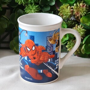 Puede incluir: Una taza de cer&aacute;mica blanca con una ilustraci&oacute;n azul, roja y amarilla de Spider-Man balance&aacute;ndose por un horizonte urbano. La taza tiene un asa y el texto "&copy;MARVEL" en la parte inferior.