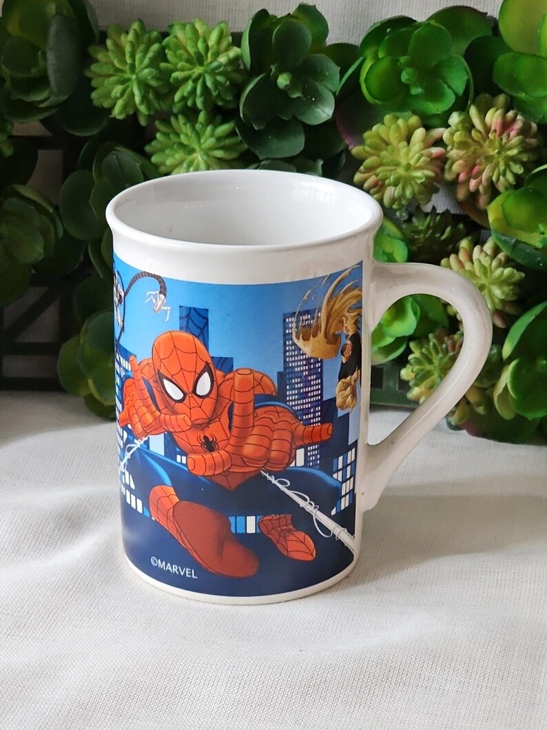 Puede incluir: Una taza de cer&aacute;mica blanca con una ilustraci&oacute;n azul, roja y amarilla de Spider-Man balance&aacute;ndose por un horizonte urbano. La taza tiene un asa y el texto "&copy;MARVEL" en la parte inferior.
