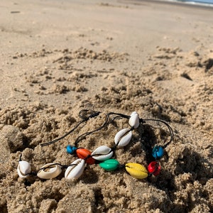 May include: A colorful shell necklace with white, orange, yellow, green, blue, and red beads on a black string. The necklace is lying on a sandy beach.