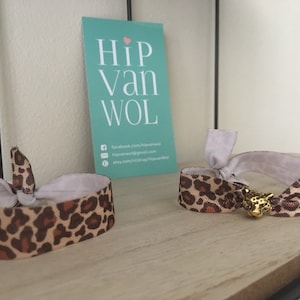 Può includere: Due scrunchies per capelli con stampa leopardata e ciondoli dorati a forma di animale. Gli scrunchies sono legati a fiocco e sono su una superficie di legno. Il testo sulla carta dietro gli scrunchies dice "Hip van Wol".