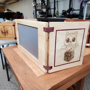 May include: A wooden game master screen with three panels. The center panel has a clear acrylic insert. The right panel has a laser-engraved image of a cartoon cat with wings sitting on a 20-sided die. The left panel has a laser-engraved design of a dragon.