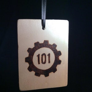 May include: A wooden tag with a brown gear design and the number 101 inside. The tag is hanging from a black ribbon.
