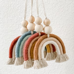 May include: A set of three rainbow-colored macrame wall hangings with wooden beads. The rainbows are made with different colored yarns and have a fringe at the bottom. The wooden beads are strung on twine and hang above the rainbows.