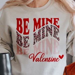 May include: A cream-colored sweatshirt with the words "BE MINE" in red, leopard print, and pink lettering. The word "Valentine" is written in red script with a small red heart. The image is a PNG digital design.