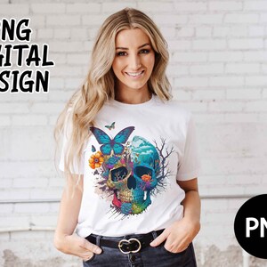 Op de afbeelding: Een wit T-shirt met een kleurrijk schedelontwerp met vlinders, bloemen en een blauw en groen landschap. De schedel is omgeven door een zwarte omtrek en heeft een witte achtergrond.