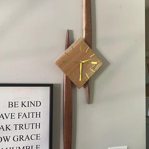 May include: A mid-century modern wooden wall clock with a diamond-shaped face and gold hands. The clock is mounted on a wall with two vertical wooden accents. The clock's face is made of light-colored wood.