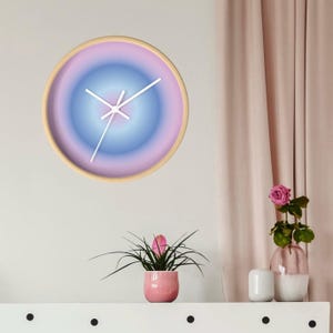 May include: A circular wall clock with a light wood frame and a gradient face that transitions from a soft pink outer edge to a deep blue center. The clock features three thin white hands positioned against the smooth, circular color gradient.