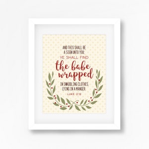 May include: A white framed print with a beige background and a green wreath with red berries. The text reads "AND THIS SHALL BE A SIGN UNTO YOU: YE SHALL FIND the babe wrapped IN SWADDLING CLOTHES, LYING IN A MANGER. LUKE 2:12"