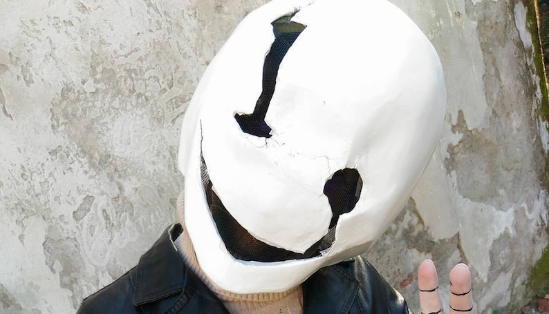 May include: A white mask with a large, toothy smile and black eye holes. The mask is made of a hard material and has a rough texture. The mask is being worn by a person, but only the mask is visible.