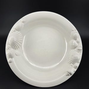 May include: A round, white ceramic plate featuring a smooth, concave center and a wide rim decorated with raised, three-dimensional seashell motifs. The relief designs include various scallop and spiral shell shapes integrated into the border.