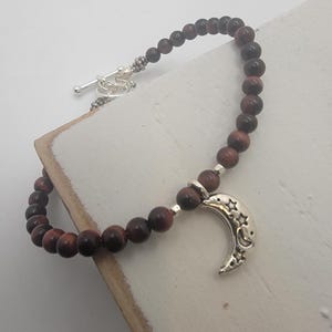 May include: A beaded bracelet featuring dark red-brown beads and a silver crescent moon charm with star cutouts. The bracelet has a silver toggle clasp. The beads are approximately 0.3 inches in diameter.