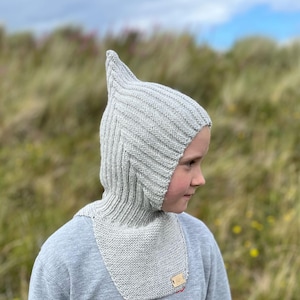 May include: A grey knitted balaclava with a pointed top, worn by a child. The balaclava covers the head and neck, leaving only the face exposed.