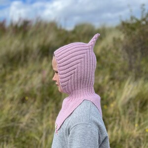May include: A pink knitted balaclava with a pointed top, worn by a person standing in a grassy field.