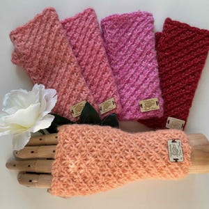 May include: Five pairs of knitted fingerless gloves in various shades of pink and red. The gloves are displayed on a wooden hand and a white rose.