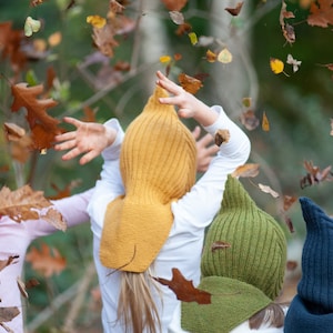 May include: Three children wearing colorful knitted hats with earflaps. The hats are yellow, green, and blue. The children are standing in a forest with autumn leaves falling around them.