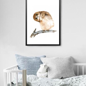 Puede incluir: Pintura a la acuarela de un b&uacute;ho curioso posado en una rama, sobre un fondo blanco dentro de un marco negro. El b&uacute;ho tiene plumas marrones y beige y ojos grandes y expresivos. La obra de arte se exhibe sobre una cuna con almohadas y un oso de peluche.