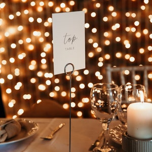 May include: A white card with the word "top table" written on it is displayed on a silver stand. The card is placed on a table with two wine glasses and a candle.