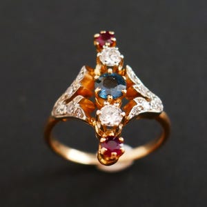 Op de afbeelding: Een gouden ring met een centrale blauwe edelsteen, geflankeerd door twee witte edelstenen en twee rode edelstenen. De ring heeft een sierlijk ontwerp met kleine witte edelstenen die in de gouden band zijn gezet.