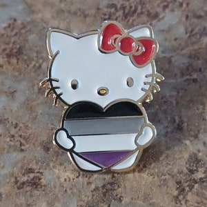 Puede incluir: Un pin de color dorado con Hello Kitty sosteniendo un símbolo en forma de corazón con rayas negras, grises, blancas y moradas. El pin tiene un lazo rojo en la cabeza de Hello Kitty.