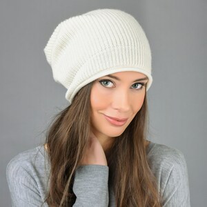 May include: A woman with long brown hair is wearing a white knit beanie and a gray sweater. She is looking at the camera with a slight smile.