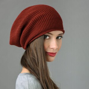 May include: A woman with long brown hair is wearing a slouchy, burgundy beanie. She is looking over her shoulder and smiling.