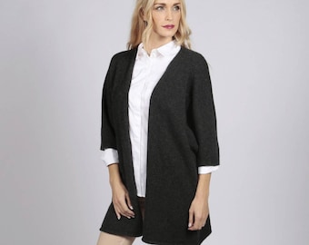 May include: A charcoal grey cardigan with a v-neck and long sleeves. The cardigan is worn over a white button-down shirt and beige trousers. The model is wearing black heels.