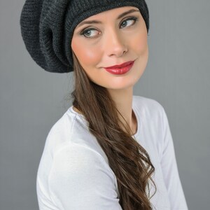 May include: A woman wearing a dark gray slouchy beanie with long brown hair. She is wearing a white long-sleeved shirt.