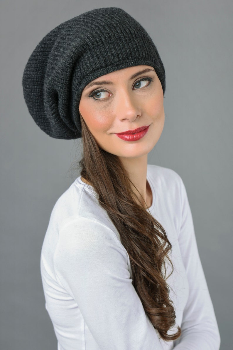 May include: A woman wearing a dark gray slouchy beanie with long brown hair. She is wearing a white long-sleeved shirt.